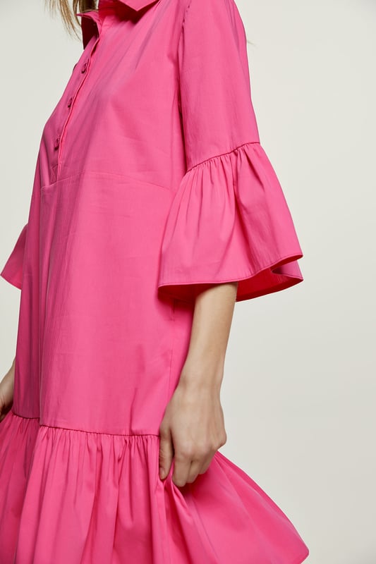 Deze fuchsia jurk is gemaakt van geweven stretch poplin. Het heeft klokkende mouwen van ¾ lengte. De jurk heeft een kraag en een sluiting in de taille met plastic knopen. Een naad onder de buste zorgt voor een perfecte pasvorm. De jurk heeft een brede zoom met ruches. De jurk heeft een losse pasvorm en valt boven de knie.