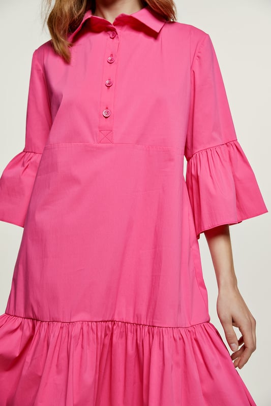 Deze fuchsia jurk is gemaakt van geweven stretch poplin. Het heeft klokkende mouwen van ¾ lengte. De jurk heeft een kraag en een sluiting in de taille met plastic knopen. Een naad onder de buste zorgt voor een perfecte pasvorm. De jurk heeft een brede zoom met ruches. De jurk heeft een losse pasvorm en valt boven de knie.