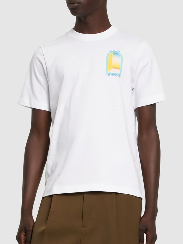 The T-shirt features a classic cut with a round neckline, offering a timeless silhouette. The front is graced with a vibrant, stylised print depicting an abstract representation of a window or gateway, bathed in the soft pastel hues of blue, yellow, and orange, evoking the serene beauty of a sunset. 