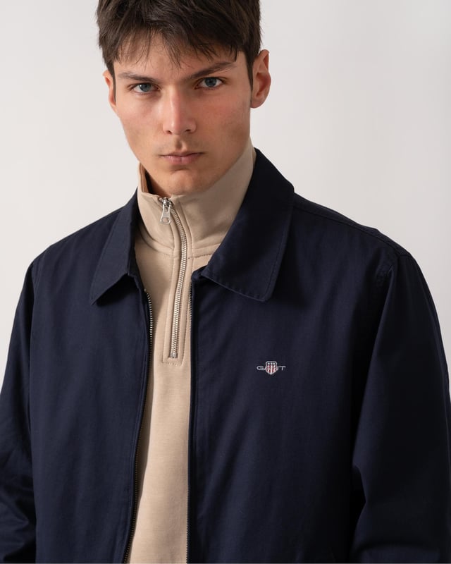 This jacket takes inspiration from GANT's beloved windcheater design, incorporating updated elements for the new season. Crafted from a water-repellent cotton twill shell, it is half-lined to shield against the cold while ensuring breathability. Designed for a regular fit that sits just above the hips, it features a partly elasticated hem for added comfort. Additional details include a front zip closure with a leather extender, slanted side welt pockets with hidden press buttons, two interior jetted pockets, and buttoned cuffs. It is finished with logo embroidery at the chest and a signature branded locker loop at the back. 

Regular fit
Above hip length
Water-repellent (DWR treatment)
Two outer and inner pockets

Shell - 100% Cotton, Upper Back Lining - 100% Cotton, Sleeve Lining - 100% Recycled Polyester 
7006494