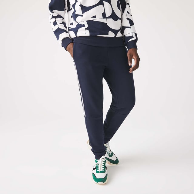 Men's Lacoste Branded Bands Skinny Fleece Joggers.- Brushed cotton blend fleece.- Elasticised waistband.- Tapered fit.- Tapered cut.- Side pockets, ribbed finishes on leg bottoms and Lacoste branding on stripes on sides.- Embroidered green crocodile on hip. Material: 83% Cotton, 17% Polyester. Ref: XH1208166.