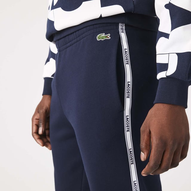 Men's Lacoste Branded Bands Skinny Fleece Joggers.- Brushed cotton blend fleece.- Elasticised waistband.- Tapered fit.- Tapered cut.- Side pockets, ribbed finishes on leg bottoms and Lacoste branding on stripes on sides.- Embroidered green crocodile on hip. Material: 83% Cotton, 17% Polyester. Ref: XH1208166.
