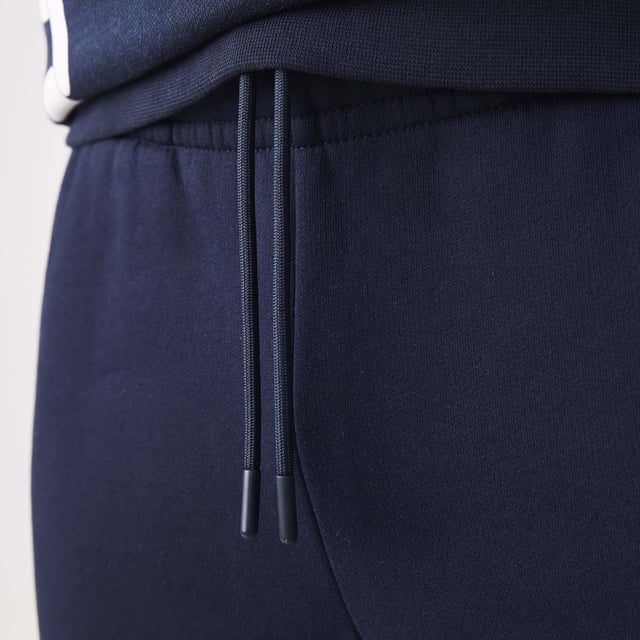 Men's Lacoste Branded Bands Skinny Fleece Joggers.- Brushed cotton blend fleece.- Elasticised waistband.- Tapered fit.- Tapered cut.- Side pockets, ribbed finishes on leg bottoms and Lacoste branding on stripes on sides.- Embroidered green crocodile on hip. Material: 83% Cotton, 17% Polyester. Ref: XH1208166.