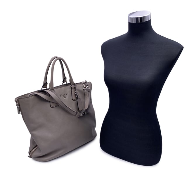 VINTAGE, UVP WIE NEU  
Schöne PRADA Tote-Bag aus strukturiertem Leder in Grau Taupe. Sie verfügt über doppelte, runde Obergriffe und einen verstellbaren sowie abnehmbaren Schultergurt. Auf der Vorderseite befindet sich die PRADA-Beschriftung in Silbermetall. Oberer Reißverschlussverschluss. 4 Fuß aus Metall am Boden. Die Tasche hat ein Hauptfach. Das Innere ist vollständig mit dem charakteristischen PRADA-Stoff ausgekleidet. 1 interne Reißverschlusstasche und 1 offene Tasche innen. Vier Metallfüße am Boden für Stil und Schutz. 'PRADA Milano'-Tag innen, kleines Etikett mit Zahlen in der Innenstasche. Zustand Bewertung: B - SEHR GUTER ZUSTAND  
Zustandsdetails: Einige kleine Kratzer (mit kleinen weißen Stellen) auf dem Leder aufgrund normaler Nutzung. Ein paar schwarze Flecken auf dem Leder durch normale Nutzung. Einige Gebrauchsspuren an den unteren Ecken. Einige leichte Kratzer auf der Oberfläche des Beschlags. Bitte überprüfen Sie die Fotos sorgfältig und fragen Sie, wenn Sie weitere Details benötigen oder zweifeln haben.  
Maße (cm):  
Tiefe: 15,2  
Höhe: 34,3  
Länge: 36,8  
Weitere Details: Staubbeutel? Ja  
Schlüssel? Nein  
Box? Nein  
Maße: Breite: 152 mm, Länge: 368 mm, Höhe: 343 mm