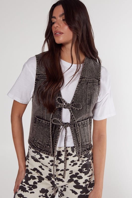 Sleeveless denim gilet with a trendy tie-front design
        Cropped length for a modern, fashion-forward silhouette
        Distinctive contrast stitching adds visual interest
        Two functional front patch pockets for practicality
        Versatile layering piece suitable for various casual looks
    
    This NastyGal tie front denim gilet is the perfect addition to your casual wardrobe, offering a blend of style and versatility. The cropped silhouette and tie-front detail create a flattering shape, while the denim fabric provides a classic, timeless appeal. Style it over a crisp white tee and pair with the trendy printed wide-leg trousers as shown for a chic daytime ensemble. For a more relaxed look, throw it over a floaty summer dress or team with high-waisted jeans and trainers. This gilet is ideal for family gatherings, weekend brunches, or casual outings with friends. Its adaptable nature allows for easy transition from day to night - simply swap your flats for ankle boots and add some statement jewellery for an effortless evening look. The denim gilet is a wardrobe staple that will see you through multiple seasons, making it a smart investment for the fashion-savvy shopper.