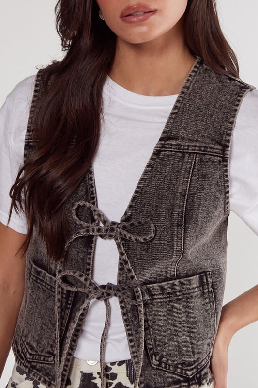 Sleeveless denim gilet with a trendy tie-front design
        Cropped length for a modern, fashion-forward silhouette
        Distinctive contrast stitching adds visual interest
        Two functional front patch pockets for practicality
        Versatile layering piece suitable for various casual looks
    
    This NastyGal tie front denim gilet is the perfect addition to your casual wardrobe, offering a blend of style and versatility. The cropped silhouette and tie-front detail create a flattering shape, while the denim fabric provides a classic, timeless appeal. Style it over a crisp white tee and pair with the trendy printed wide-leg trousers as shown for a chic daytime ensemble. For a more relaxed look, throw it over a floaty summer dress or team with high-waisted jeans and trainers. This gilet is ideal for family gatherings, weekend brunches, or casual outings with friends. Its adaptable nature allows for easy transition from day to night - simply swap your flats for ankle boots and add some statement jewellery for an effortless evening look. The denim gilet is a wardrobe staple that will see you through multiple seasons, making it a smart investment for the fashion-savvy shopper.