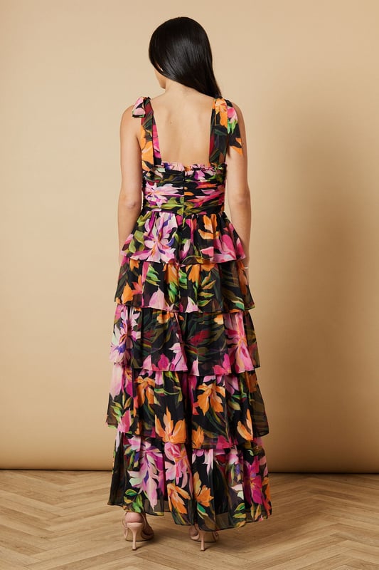 Feminine ruched bodice with elegant gathering that creates a flattering silhouette
        Charming tie straps with ruffle detail for an adjustable fit and pretty finish
        Tiered midi skirt design with beautiful movement and volume
        Striking floral print that captures Oasis's signature feminine aesthetic
        Square neckline that frames the dcolletage beautifully
    
    This enchanting midi dress from Oasis embodies romantic charm with its thoughtfully designed ruched bodice and playful tiered skirt. The delicate tie straps can be adjusted to your perfect fit, while the ruffle detailing adds a touch of whimsy to this occasion-ready piece. Style with strappy heeled sandals as seen on our model for garden parties and summer weddings, or dress it up with statement earrings and an elegant clutch for formal events. The flowing tiered design creates beautiful movement as you walk, making it ideal for those special moments that call for something truly memorable. For cooler evenings, layer with a lightweight pashmina or tailored blazer to maintain that refined silhouette whilst staying comfortable throughout your event.