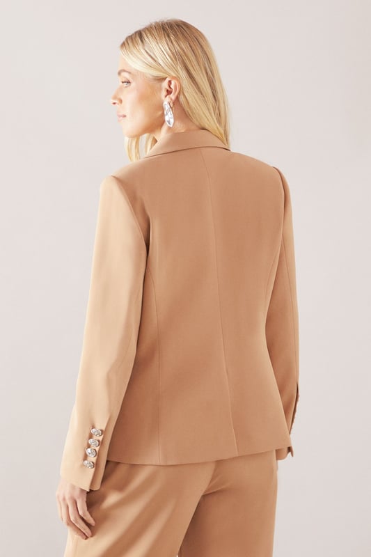 Double-breasted design with striking metallic button detailing for a sophisticated silhouette
        Tailored notched lapels that frame the face beautifully
        Structured shoulders creating a refined, feminine shape
        Functional front flap pockets adding practical elegance
        Decorative sleeve buttons that enhance the tailored finish
    
    This petite double-breasted blazer offers the perfect balance of sophistication and contemporary style. Designed specifically for smaller frames, it creates a flattering silhouette without overwhelming your proportions. The structured shoulders and defined waist work harmoniously to create a polished look suitable for both professional settings and formal occasions. Style with the matching trousers as seen on our model for a coordinated suit ensemble, or pair with slim-fit trousers and heels for a business meeting. For evening events, layer over a silk camisole and add statement jewellery like the drop earrings shown. The versatile design transitions seamlessly from day to night, making it an essential wardrobe investment for the woman who appreciates timeless glamour with modern sensibility.