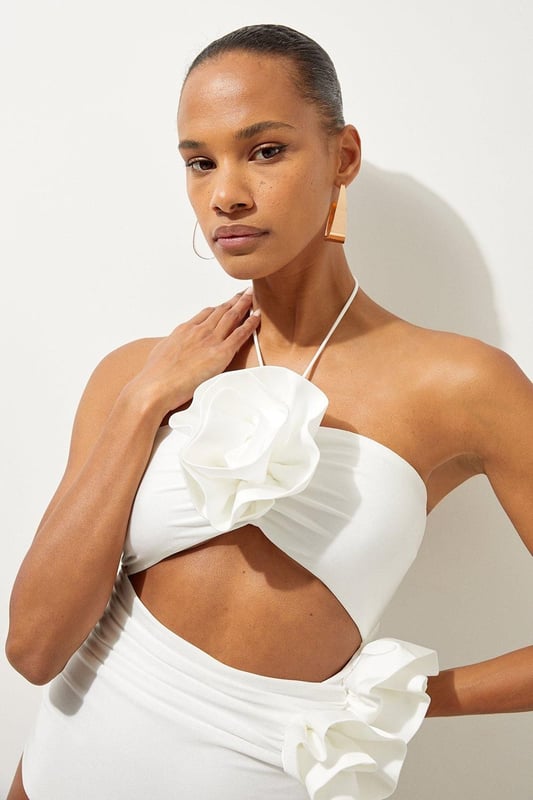 Welcome the sun with a swimming costume that will turn heads, featuring a three dimensional corsage accent on the bust and hip, and a statement cut out design in the front and back. Style this piece with strappy sandals and a beach cover up for a holiday look that can be reworn time and time again. Rosette Cut Out Detail Swimsuit Flattering Halter Neckline Unique, Three Dimensional Flower Accent Statement Cut Out Design Tie and Clasp Fastening in Back
