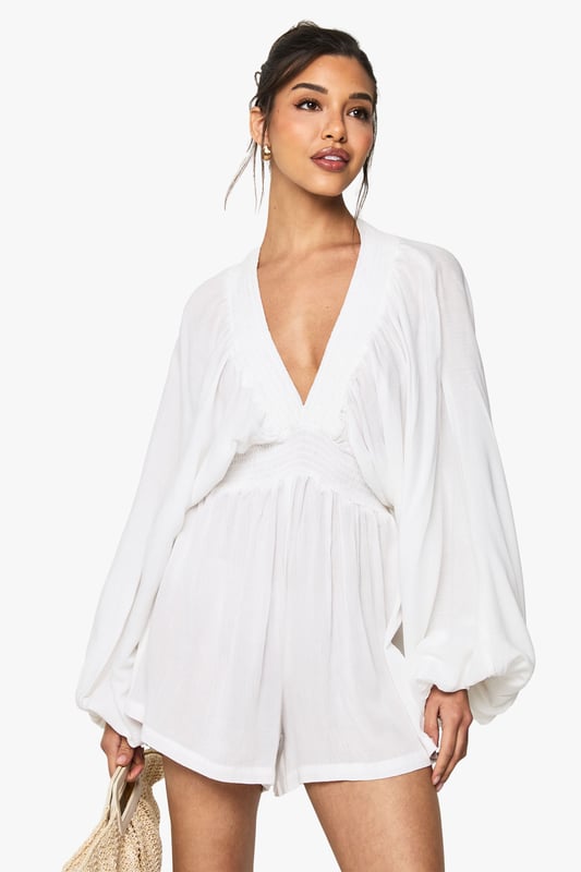Lightweight cheesecloth playsuit with plunge neckline for a striking silhouette
        Dramatic long sleeves with voluminous design for added flair
        Elasticated waistband creates a flattering, defined shape
        Relaxed fit shorts provide comfort and ease of movement
        Back keyhole detail with button fastening adds a subtle finishing touch
    
    This effortlessly chic playsuit from Boohoo combines comfort with head-turning style. The lightweight cheesecloth fabric drapes beautifully across the body, while the plunging neckline creates a bold focal point that's perfect for warm-weather socialising. The billowing long sleeves add drama to this casual piece, balancing the shorter length of the playsuit. Style with strappy sandals and a woven clutch as seen on our model for daytime gatherings, or dress up with metallic accessories and statement earrings for sunset drinks. The versatile design works brilliantly for family barbecues, beach outings or casual coffee dates. For cooler evenings, layer with a denim jacket and switch to ankle boots for an outfit that transitions seamlessly from day to night. Machine washable and easy to care for, this playsuit will quickly become your go-to for effortless summer style.