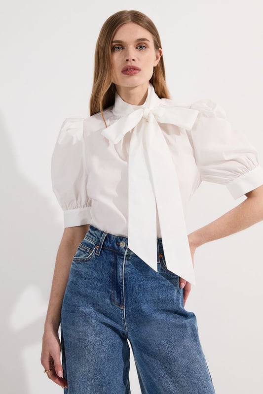 This Karen Millen blouse artfully balances classic femininity with contemporary design elements. The statement pussy bow and dramatic puff sleeves create a silhouette that commands attention whilst maintaining effortless sophistication. Style with high-waisted denim for casual weekend brunches, as seen on our model, or pair with tailored trousers for polished daywear options. The structured cotton poplin fabric holds its shape beautifully throughout the day, making this piece ideal for family gatherings or important daytime meetings. The unexpected back cut-out detail adds interest when viewed from all angles, ensuring you make an impression coming and going. This versatile blouse transitions seamlessly from day to evening; simply swap your trainers for a block heel and add statement jewellery for dinner dates or gallery openings.  Elegant pussy bow detail creates a sophisticated focal point Voluminous puff sleeves add a contemporary silhouette High neckline offers refined coverage for versatile styling Crisp cotton poplin fabric provides structure with breathable comfort Distinctive cut-out back detail introduces an unexpected modern twist