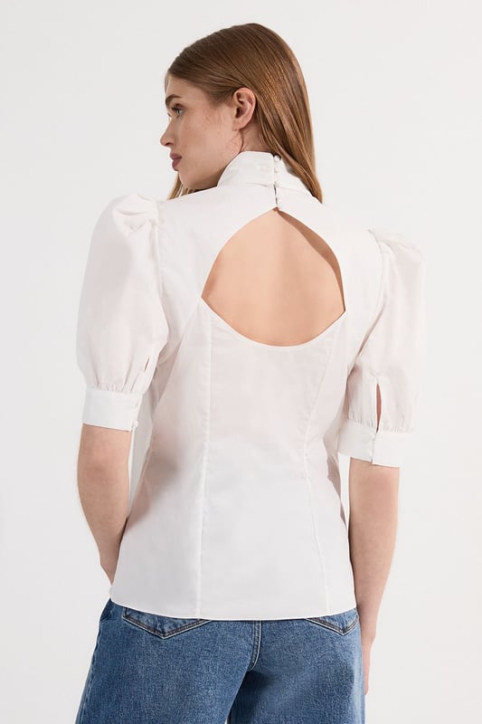 This Karen Millen blouse artfully balances classic femininity with contemporary design elements. The statement pussy bow and dramatic puff sleeves create a silhouette that commands attention whilst maintaining effortless sophistication. Style with high-waisted denim for casual weekend brunches, as seen on our model, or pair with tailored trousers for polished daywear options. The structured cotton poplin fabric holds its shape beautifully throughout the day, making this piece ideal for family gatherings or important daytime meetings. The unexpected back cut-out detail adds interest when viewed from all angles, ensuring you make an impression coming and going. This versatile blouse transitions seamlessly from day to evening; simply swap your trainers for a block heel and add statement jewellery for dinner dates or gallery openings.  Elegant pussy bow detail creates a sophisticated focal point Voluminous puff sleeves add a contemporary silhouette High neckline offers refined coverage for versatile styling Crisp cotton poplin fabric provides structure with breathable comfort Distinctive cut-out back detail introduces an unexpected modern twist