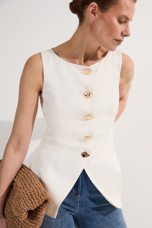 Ease into the new season, surrendering to the return of sunlit afternoons and a relaxed, carefree spirit. Discover contemporary cuts that can be mixed and matched effortlessly - creating a clean and sophisticated aesthetic.  This waistcoat reimagines traditional tailoring with its contemporary silhouette and striking button detail. The linen-viscose blend creates a structured yet breathable piece that maintains its shape beautifully throughout the day. Style with wide-leg trousers as shown for a relaxed daytime ensemble that balances structure with fluidity. For casual gatherings, layer over a crisp white shirt with sleeves rolled up and pair with your favourite straight-leg jeans and leather trainers. This versatile piece transitions effortlessly from office to weekend brunch, simply swap your footwear from flats to ankle boots as the occasion demands. The unique button placement and asymmetrical hem create a distinctive focal point that will refresh your everyday wardrobe staples.  Sleek tailored waistcoat featuring distinctive statement buttons for a contemporary twist Slim fit design with a flattering silhouette that accentuates the waistline Round neckline offering a sophisticated alternative to traditional V-neck waistcoats Asymmetrical hem with a modern front split detail for added visual interest Versatile sleeveless design perfect for layering throughout multiple seasons