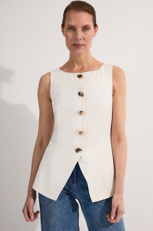Ease into the new season, surrendering to the return of sunlit afternoons and a relaxed, carefree spirit. Discover contemporary cuts that can be mixed and matched effortlessly - creating a clean and sophisticated aesthetic.  This waistcoat reimagines traditional tailoring with its contemporary silhouette and striking button detail. The linen-viscose blend creates a structured yet breathable piece that maintains its shape beautifully throughout the day. Style with wide-leg trousers as shown for a relaxed daytime ensemble that balances structure with fluidity. For casual gatherings, layer over a crisp white shirt with sleeves rolled up and pair with your favourite straight-leg jeans and leather trainers. This versatile piece transitions effortlessly from office to weekend brunch, simply swap your footwear from flats to ankle boots as the occasion demands. The unique button placement and asymmetrical hem create a distinctive focal point that will refresh your everyday wardrobe staples.  Sleek tailored waistcoat featuring distinctive statement buttons for a contemporary twist Slim fit design with a flattering silhouette that accentuates the waistline Round neckline offering a sophisticated alternative to traditional V-neck waistcoats Asymmetrical hem with a modern front split detail for added visual interest Versatile sleeveless design perfect for layering throughout multiple seasons