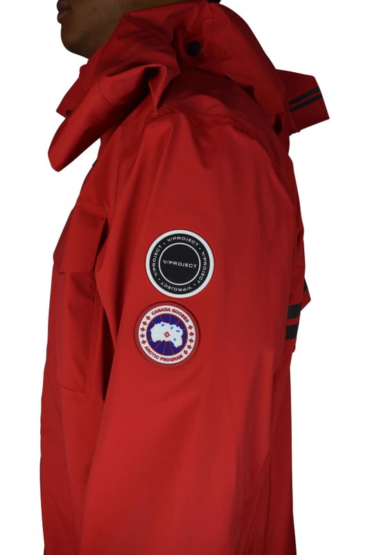 Rote Jacke im Stil von K-Way Y/Project Canada Goose.Stehkragen mit Kapuze.Reißverschluss mit Druckknöpfen.Lange Ärmel.Reflektierende Streifen auf der Rückseite.Aufgenähte Logo-Patches auf dem linken Ärmel.Klettverschlussstreifen zum Verengen an den Ärmeln.Zwei Seitentaschen mit Reißverschluss.Tasche mit Klettverschluss auf der linken Brust.