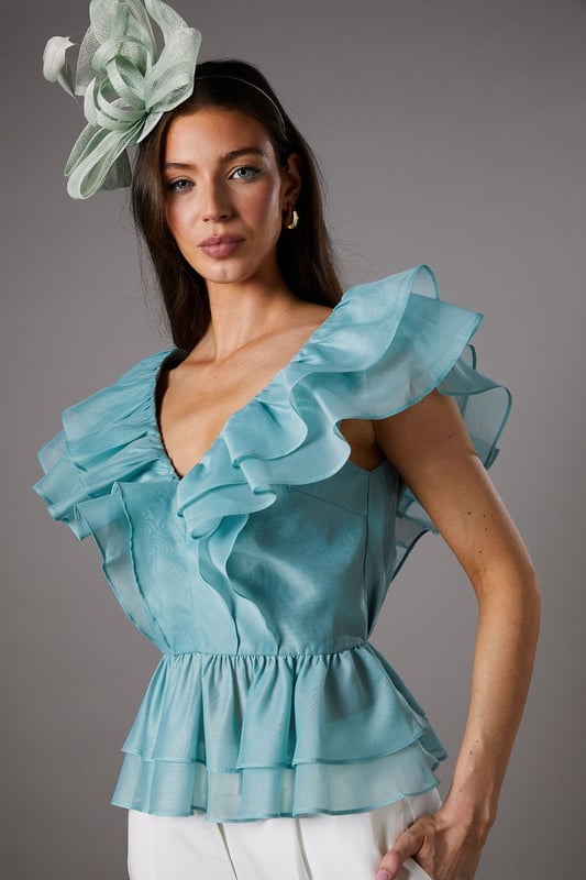 Exquisite organza fabric with delicate texture for a refined finish
        Dramatic V-neckline complemented by cascading frills for a feminine silhouette
        Elegant peplum design creating a flattering waistline definition
        Striking open back with ruffle detailing for added drama
        Tiered hem with multiple layers of frills for dimensional movement
    
    This captivating Coast peplum top showcases the beauty of textured organza with its sumptuous frills and feminine detailing. The striking V-neckline frames the dcolletage beautifully, whilst the peplum silhouette creates a figure-enhancing shape that celebrates the waistline. Perfect for summer race days or wedding celebrations, this top pairs wonderfully with tailored wide-leg trousers as styled on our model. Add a statement fascinator and minimal jewellery to complete your occasion ensemble. For evening events, swap the fascinator for chandelier earrings and a sleek clutch bag. The versatile design works brilliantly for garden parties, formal luncheons or champagne receptions where sophisticated dressing is essential. The open back detail adds an unexpected touch of allure to this otherwise classic piece.