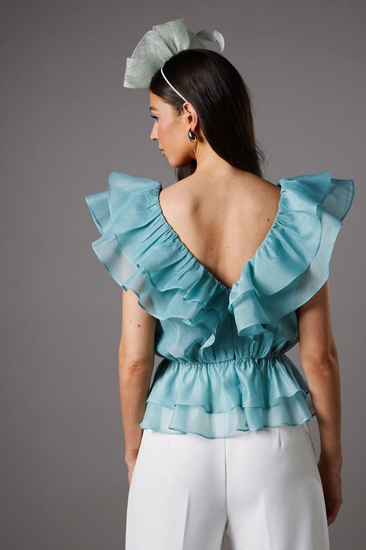 Exquisite organza fabric with delicate texture for a refined finish
        Dramatic V-neckline complemented by cascading frills for a feminine silhouette
        Elegant peplum design creating a flattering waistline definition
        Striking open back with ruffle detailing for added drama
        Tiered hem with multiple layers of frills for dimensional movement
    
    This captivating Coast peplum top showcases the beauty of textured organza with its sumptuous frills and feminine detailing. The striking V-neckline frames the dcolletage beautifully, whilst the peplum silhouette creates a figure-enhancing shape that celebrates the waistline. Perfect for summer race days or wedding celebrations, this top pairs wonderfully with tailored wide-leg trousers as styled on our model. Add a statement fascinator and minimal jewellery to complete your occasion ensemble. For evening events, swap the fascinator for chandelier earrings and a sleek clutch bag. The versatile design works brilliantly for garden parties, formal luncheons or champagne receptions where sophisticated dressing is essential. The open back detail adds an unexpected touch of allure to this otherwise classic piece.