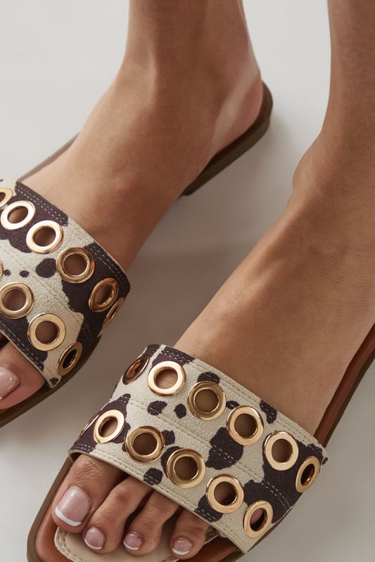 Faux suede sliders featuring striking cow print pattern for a bold statement
        Distinctive metallic eyelet detailing across the wide strap adds contemporary edge
        Comfortable flat heel measuring between 0-2cm for all-day wearability
        Open-toe design with slip-on style for effortless summer dressing
        Cushioned footbed provides essential comfort for casual everyday wear
    
    These Faith Richie sliders combine eye-catching design with practical comfort for your warm-weather wardrobe. The standout cow print and metallic eyelet detailing transform a casual footwear essential into a fashion statement piece. Style these versatile sliders with denim shorts and a simple white tee for relaxed weekend brunches, or pair with a floaty summer dress for garden parties. The flat heel ensures comfort whilst strolling through farmers' markets or enjoying casual family gatherings. For beach holidays, these sliders transition effortlessly from poolside to beachfront caf. Their slip-on design makes them perfect for busy mornings when you need to dash out the door without compromising on style. The neutral tones within the pattern ensure these sliders will complement numerous outfits in your summer collection.