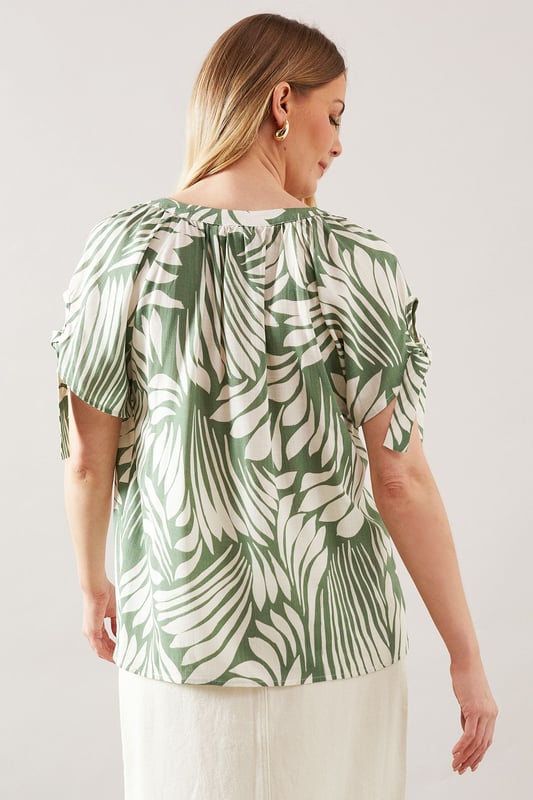 Distinctive collarless design with elegant V-neckline for a sophisticated silhouette
        Unique tie sleeve detail adding a touch of feminine charm to this woven top
        Relaxed fit through the body for all-day comfort and effortless movement
        Versatile abstract print that makes a statement whilst remaining wonderfully wearable
        Short sleeves with decorative ties that create a graceful drape effect
    
    This captivating collarless top from Wallis combines comfort with sophisticated style. The eye-catching abstract print creates visual interest whilst the tie sleeve detail adds a touch of feminine flair to your daywear collection. Style with a belted cream midi skirt and leather sandals for a refined lunch date look, or pair with tailored trousers and ballet flats for family gatherings. The V-neckline frames the dcolletage beautifully, making it perfect for showcasing delicate jewellery. The viscose-blend fabric offers a lovely drape that flatters the figure without clinging. This versatile piece transitions seamlessly from casual coffee mornings to afternoon garden parties, making it an essential addition to your wardrobe that works across multiple seasons.