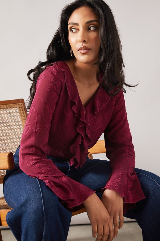 Elegant jacquard fabric with subtle textured pattern throughout
        Feminine ruffle detail cascading down the front
        V-neckline framed by delicate ruffles for a sophisticated touch
        Flared bell sleeves with ruffle trim for added movement
        Relaxed silhouette perfect for petite frames
    
    This charming Wallis blouse combines sophistication with everyday wearability. The jacquard fabric presents a subtle textured pattern that catches the light beautifully, whilst the cascading ruffle detail adds a feminine flourish to your daytime ensemble. Style with your favourite denim jeans and white trainers for casual family gatherings, or tuck into tailored trousers with ballet flats for lunch with friends. The bell sleeves create elegant movement as you go about your day, making this piece both practical and pretty. Layer under a cardigan when the temperature drops or wear alone with statement earrings to showcase the beautiful neckline detail. This versatile blouse transitions effortlessly from shopping trips to informal dinners, making it a wardrobe essential for the sophisticated woman who values both comfort and style.