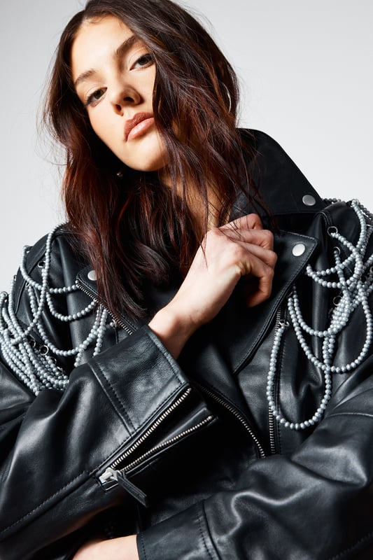 Genuine leather jacket with striking pearl trim detailing draped across the shoulders and back
        Classic biker silhouette with notched lapels and asymmetric zip fastening
        Decorative fringe trim cascading from the pearl embellishments for dramatic movement
        Functional side pockets and adjustable buckle detail at the hem
        Fully lined for comfort and ease of wear
    
    Make a bold statement with this Nasty Gal leather jacket that reimagines the classic biker style with eye-catching pearl trim detailing. The dramatic draping pearl chains and fringe create a captivating silhouette that demands attention, perfect for those who aren't afraid to stand out. Style it as seen with a floaty white mini dress and western-inspired boots for an edgy contrast that works brilliantly for nights out. For a more structured evening look, pair with a fitted bodycon dress and strappy heels. The versatile design allows for both rebellious and refined styling options, making it an ideal choice for everything from gigs and club nights to cocktail bars. This jacket doesn't just complete an outfit-it defines it, bringing undeniable character to your night-out wardrobe.