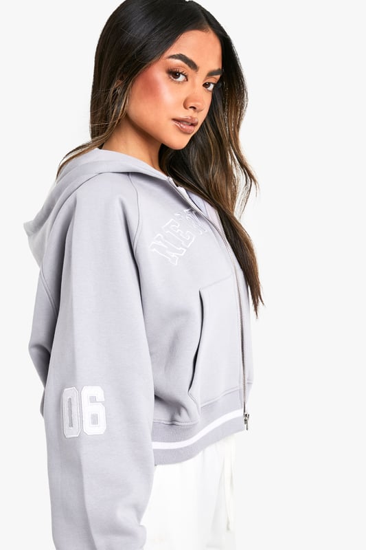 Cropped design for a trendy silhouette
        Full-length zip closure for versatile styling
        Bold 'New York' slogan across the chest
        '06' detail on the sleeve for a sporty touch
        Comfortable hood for added warmth and style
    
    Make a statement with this eye-catching cropped hoodie, perfect for the fashion-forward trendsetter. The bold 'New York' slogan and '06' detail give it an urban, street-style edge that's sure to turn heads. Its versatile design allows for effortless styling - pair it with high-waisted trousers and trainers for a casual daytime look, or throw it over a bodycon dress for an edgy night out ensemble. The full-length zip offers adaptability, allowing you to customise your look to suit the occasion or weather. Ideal for everything from coffee dates to weekend shopping trips, this hoodie is a must-have addition to any casual wardrobe. Its cosy hood and soft fabric ensure comfort without compromising on style, making it the perfect go-to piece for those days when you want to look effortlessly cool.