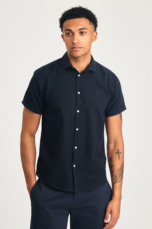 This short sleeve cotton-linen mix shirt by Nines features a sleek embroidered logo on the chest. Lightweight and breathable, it’s perfect for warm days. A stylish and comfortable addition to your wardrobe, offering the ideal balance of relaxed yet refined style.