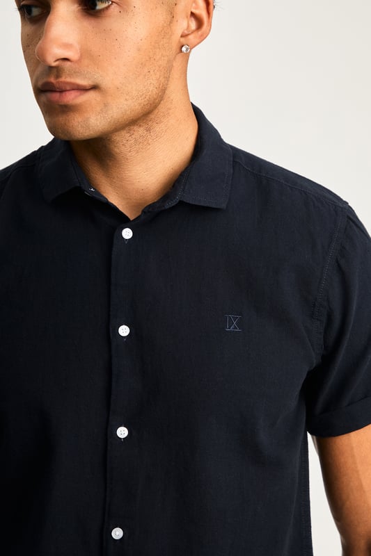 This short sleeve cotton-linen mix shirt by Nines features a sleek embroidered logo on the chest. Lightweight and breathable, it’s perfect for warm days. A stylish and comfortable addition to your wardrobe, offering the ideal balance of relaxed yet refined style.
