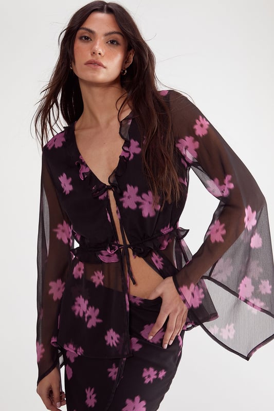Lightweight chiffon fabric with all-over ditsy floral print
        Elegant tie-front closure with ruffle detailing
        Dramatic fluted sleeves for statement appeal
        Feminine V-neckline with subtle ruffle trim
        Semi-sheer finish for layered styling versatility
    
    This enchanting Nasty Gal top showcases the perfect balance between bohemian flair and contemporary chic. The delicate chiffon construction creates a floaty silhouette, while the distinctive fluted sleeves add dramatic movement to your ensemble. Style with high-waisted trousers and strappy heels for an evening soire, or pair with your favourite denim and ankle boots for a more casual approach. The tie-front detail allows for adjustable styling, making this piece wonderfully adaptable for various occasions. For office wear, layer with a fitted camisole underneath and team with tailored trousers for a look that transitions seamlessly from desk to dinner. The ruffle detailing adds a touch of romance, perfect for date nights or special gatherings where you want to make a subtle yet sophisticated statement.