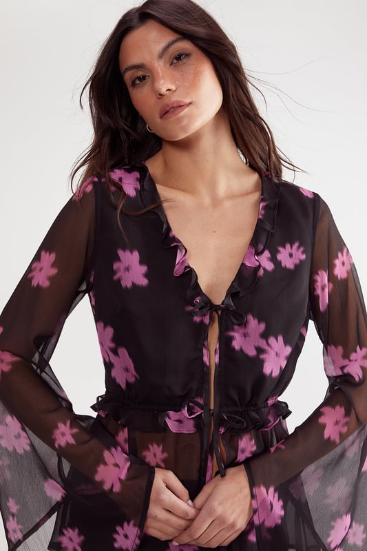Lightweight chiffon fabric with all-over ditsy floral print
        Elegant tie-front closure with ruffle detailing
        Dramatic fluted sleeves for statement appeal
        Feminine V-neckline with subtle ruffle trim
        Semi-sheer finish for layered styling versatility
    
    This enchanting Nasty Gal top showcases the perfect balance between bohemian flair and contemporary chic. The delicate chiffon construction creates a floaty silhouette, while the distinctive fluted sleeves add dramatic movement to your ensemble. Style with high-waisted trousers and strappy heels for an evening soire, or pair with your favourite denim and ankle boots for a more casual approach. The tie-front detail allows for adjustable styling, making this piece wonderfully adaptable for various occasions. For office wear, layer with a fitted camisole underneath and team with tailored trousers for a look that transitions seamlessly from desk to dinner. The ruffle detailing adds a touch of romance, perfect for date nights or special gatherings where you want to make a subtle yet sophisticated statement.