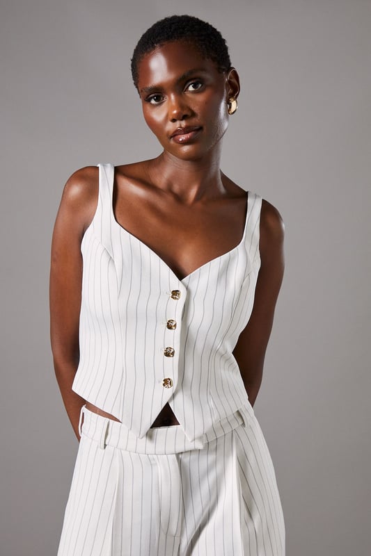 Elegant strappy waistcoat featuring a sophisticated pinstripe pattern
        Flattering V-neckline complemented by wide shoulder straps for comfort
        Decorative button-through front with contrasting metallic buttons
        Tailored fit with a slight stretch for a figure-enhancing silhouette
        Versatile cropped length perfect for pairing with high-waisted bottoms
    
    This refined Coast waistcoat brings contemporary sophistication to occasion dressing. The classic pinstripe pattern is reimagined with a feminine twist through the strappy design and fitted silhouette. Style with the matching wide-leg trousers as seen on our model for an impeccably coordinated ensemble that commands attention at summer weddings or race day events. For evening occasions, pair with a sleek pencil skirt and delicate jewellery for a look that conveys understated elegance. The crepe fabric offers comfortable wear throughout the day while maintaining its structured shape. This versatile piece transitions beautifully from formal daytime events to evening soires with just a change of accessories - try statement earrings and a clutch bag to transform your look as day turns to night.