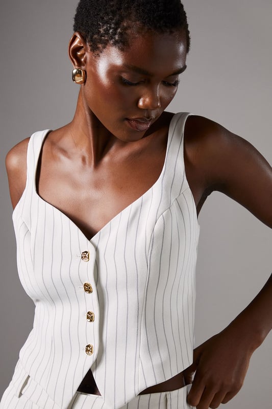 Elegant strappy waistcoat featuring a sophisticated pinstripe pattern
        Flattering V-neckline complemented by wide shoulder straps for comfort
        Decorative button-through front with contrasting metallic buttons
        Tailored fit with a slight stretch for a figure-enhancing silhouette
        Versatile cropped length perfect for pairing with high-waisted bottoms
    
    This refined Coast waistcoat brings contemporary sophistication to occasion dressing. The classic pinstripe pattern is reimagined with a feminine twist through the strappy design and fitted silhouette. Style with the matching wide-leg trousers as seen on our model for an impeccably coordinated ensemble that commands attention at summer weddings or race day events. For evening occasions, pair with a sleek pencil skirt and delicate jewellery for a look that conveys understated elegance. The crepe fabric offers comfortable wear throughout the day while maintaining its structured shape. This versatile piece transitions beautifully from formal daytime events to evening soires with just a change of accessories - try statement earrings and a clutch bag to transform your look as day turns to night.