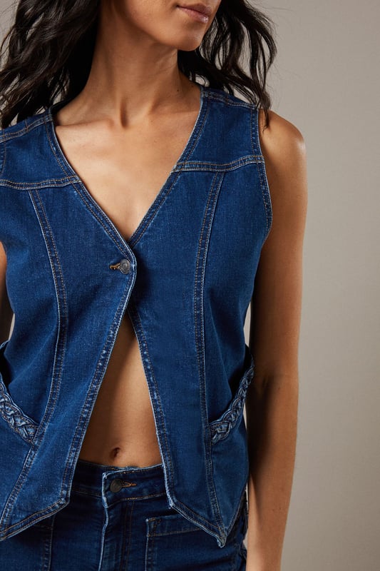 Boxy fit denim waistcoat with distinctive button front closure
        Sleeveless design with V-neckline for a feminine silhouette
        Contrast topstitching detail highlighting the structured seams
        Practical front pockets with decorative stitching
        Versatile co-ord piece that can be styled separately or as a set
    
    This Button Front Denim Waistcoat from Warehouse offers effortless edge to your casual wardrobe. The boxy silhouette creates a contemporary shape that pairs brilliantly with the matching flared trousers as seen on our model, or can be styled with your favourite denim jeans for a double denim moment. For daytime outings, layer over a simple white tee and finish with trainers and statement jewellery. When the evening calls, wear it unbuttoned over a fitted bodysuit and team with heeled boots for a touch of after-dark confidence. Perfect for festivals, weekend brunches or casual gatherings, this versatile waistcoat transitions seamlessly through the seasons - simply add layers underneath when temperatures drop. The cotton fabric ensures comfort whilst maintaining that structured denim look that never goes out of style.