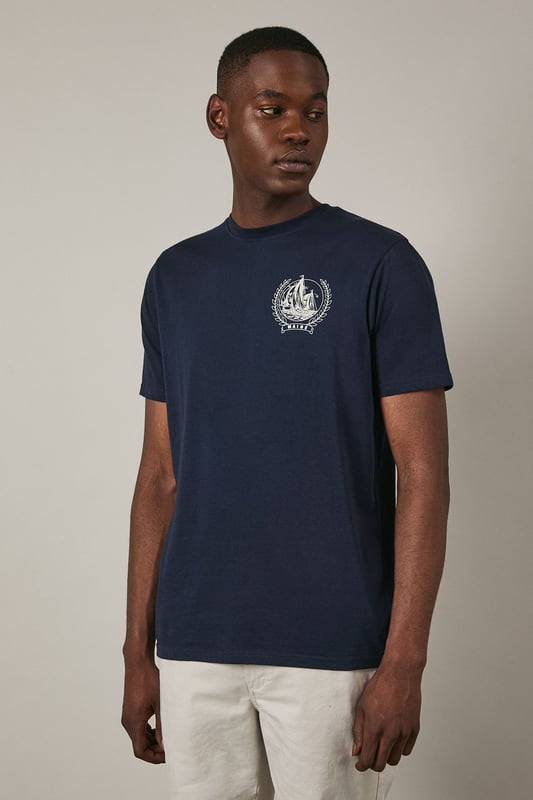 Soft jersey fabric for all-day comfort and breathability
        Classic crew neckline with ribbed trim for a neat finish
        Distinctive coastal ship emblem on chest for nautical appeal
        Short sleeves with clean-cut edges for a streamlined look
        Relaxed fit through the body offering ease of movement
    
    This Maine coastal print t-shirt brings maritime charm to your casual wardrobe. The understated design features a distinctive sailing ship emblem encircled by a laurel wreath, creating a subtle focal point against the navy background. Style it as shown with light-coloured shorts and white trainers for a fresh summer look perfect for beachside strolls or weekend brunches. For cooler evenings, layer under an unstructured blazer or light cotton overshirt. The versatile design transitions effortlessly from daytime casual to relaxed evening gatherings. The cotton jersey construction ensures this t-shirt remains comfortable throughout the day, making it an ideal choice for family outings, casual meet-ups with friends, or simply lounging at home with understated style.