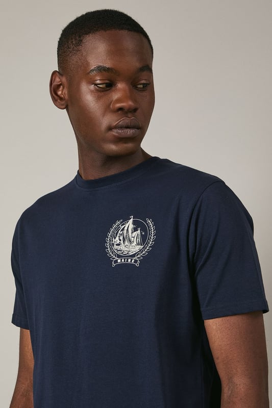 Soft jersey fabric for all-day comfort and breathability
        Classic crew neckline with ribbed trim for a neat finish
        Distinctive coastal ship emblem on chest for nautical appeal
        Short sleeves with clean-cut edges for a streamlined look
        Relaxed fit through the body offering ease of movement
    
    This Maine coastal print t-shirt brings maritime charm to your casual wardrobe. The understated design features a distinctive sailing ship emblem encircled by a laurel wreath, creating a subtle focal point against the navy background. Style it as shown with light-coloured shorts and white trainers for a fresh summer look perfect for beachside strolls or weekend brunches. For cooler evenings, layer under an unstructured blazer or light cotton overshirt. The versatile design transitions effortlessly from daytime casual to relaxed evening gatherings. The cotton jersey construction ensures this t-shirt remains comfortable throughout the day, making it an ideal choice for family outings, casual meet-ups with friends, or simply lounging at home with understated style.