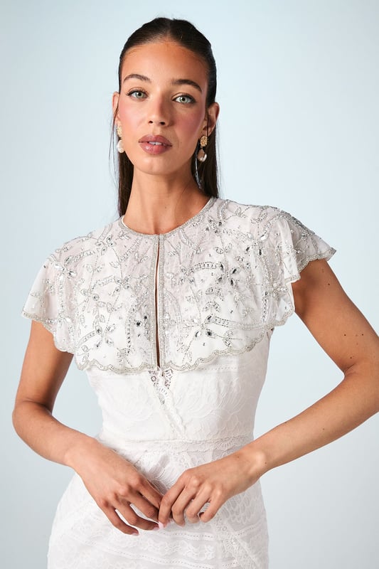 Exquisite hand-stitched cape adorned with sparkling gem embellishments and delicate sequin detailing
        Elegant flutter cap sleeves create a graceful silhouette that frames the shoulders beautifully
        Stunning open back design with sheer panels adds a contemporary touch to this traditional bridal accessory
        Intricate beadwork catches the light with every movement for a truly magical effect
        Versatile piece that can be styled with various wedding gown silhouettes for a personalised bridal look
    
    Make a breathtaking entrance on your special day with this enchanting bridal cape from Coast. The delicate hand-stitched detailing and carefully placed gem embellishments create a halo of radiance around your shoulders, adding a touch of glamour to your wedding ensemble. The thoughtfully designed open back ensures your gown remains the centrepiece whilst the cape adds dimension and visual interest. Style with a sleek updo and drop earrings as seen on our model to showcase the intricate neckline, or wear with flowing locks for a more romantic aesthetic. Perfect for the ceremony and photographs, this cape transitions beautifully from the aisle to the reception, offering a sophisticated layer that can be removed later to reveal a second look for evening celebrations. A truly special accessory that will become a cherished keepsake of your wedding day.