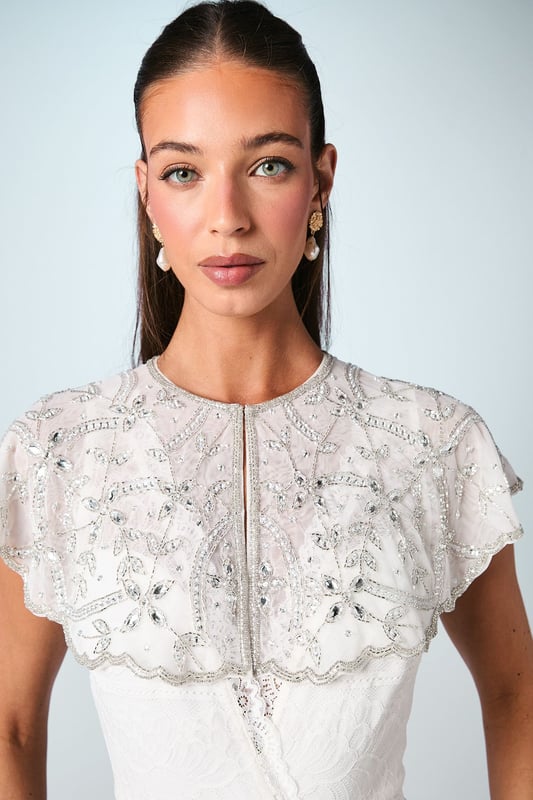 Exquisite hand-stitched cape adorned with sparkling gem embellishments and delicate sequin detailing
        Elegant flutter cap sleeves create a graceful silhouette that frames the shoulders beautifully
        Stunning open back design with sheer panels adds a contemporary touch to this traditional bridal accessory
        Intricate beadwork catches the light with every movement for a truly magical effect
        Versatile piece that can be styled with various wedding gown silhouettes for a personalised bridal look
    
    Make a breathtaking entrance on your special day with this enchanting bridal cape from Coast. The delicate hand-stitched detailing and carefully placed gem embellishments create a halo of radiance around your shoulders, adding a touch of glamour to your wedding ensemble. The thoughtfully designed open back ensures your gown remains the centrepiece whilst the cape adds dimension and visual interest. Style with a sleek updo and drop earrings as seen on our model to showcase the intricate neckline, or wear with flowing locks for a more romantic aesthetic. Perfect for the ceremony and photographs, this cape transitions beautifully from the aisle to the reception, offering a sophisticated layer that can be removed later to reveal a second look for evening celebrations. A truly special accessory that will become a cherished keepsake of your wedding day.