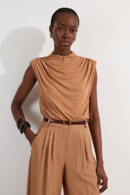This versatile Karen Millen bodysuit combines contemporary design with everyday wearability. The high neck and artful ruching create a refined silhouette that works beautifully with both casual and smart-casual ensembles. Style with high-waisted trousers as shown for a polished daytime look, perfect for brunches or gallery visits. The sleek design pairs wonderfully with wide-leg jeans and trainers for weekend outings, or dress it up with a tailored blazer and statement jewellery for dinner dates. The bodysuit format eliminates the need for constant readjustment, making it an ideal foundation piece for busy days when comfort and style must work in harmony. Add a leather jacket and ankle boots to transition this piece effortlessly into evening drinks with friends.  Elegant high neck design with sophisticated ruched detailing for a flattering silhouette Sleeveless cut with gentle draping across the bodice for effortless style Bodysuit construction ensures a smooth, tucked-in finish that stays in place all day Soft jersey crepe fabric offers comfortable stretch and movement Stylish back opening with button fastening adds an unexpected design element