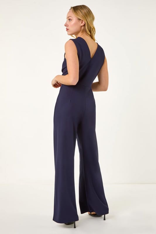 Dusk V-Neck Front Knot Jumpsuit from Roman. Be the epitome of chic in this knot jumpsuit, perfect for those warm Spring/Summer days. The flattering V-neck design with a stylish knot front detail creates a sophisticated look. This sleek sleeveless jumpsuit is a versatile piece that can be dressed up with strappy heels and a blazer for a night out or special occasion. Elevate your ensemble effortlessly and step out in style with this fashion-forward outfit!