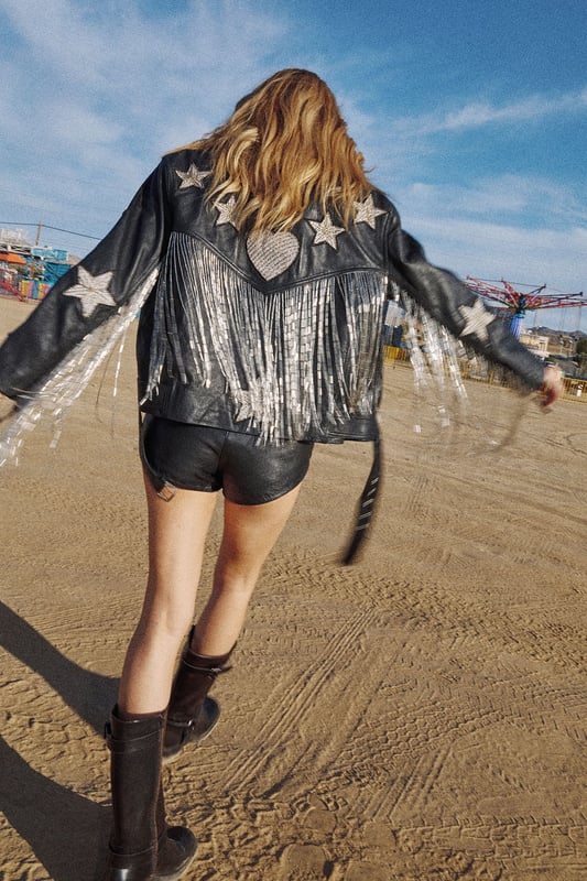 Genuine leather jacket featuring playful star and heart-shaped patches for distinctive character
        Statement metallic tassels cascade down the back for dramatic movement and flair
        Boxy, oversized silhouette offers a relaxed fit perfect for layering
        Classic biker-style design with notched lapels and asymmetric zip closure
        Fully lined interior ensures comfort and durability for long-lasting wear
    
    Make a bold statement with this eye-catching leather jacket from Nasty Gal. The oversized, boxy fit creates a contemporary silhouette that pairs brilliantly with both casual and evening looks. The striking tassel detail adds movement and drama to your ensemble, whilst the whimsical star and heart patches infuse a touch of playfulness into this edgy piece. Style with shorts and a crop top as shown for festival-ready vibes, or throw over a slip dress with chunky boots for an unforgettable night out look. Perfect for those who aren't afraid to stand out, this jacket works brilliantly for club nights, drinks with friends, or any urban adventure where you want to showcase your distinctive style. The versatile design allows for year-round wear - layer over knitwear during cooler months or drape over your shoulders on chilly summer evenings.