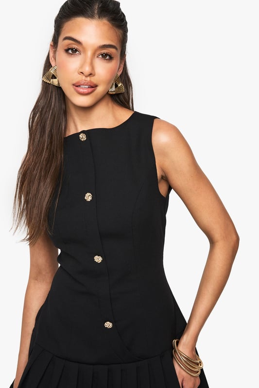 Sleeveless mini dress with elegant slash neckline for a sophisticated silhouette
        Decorative button detail running down the front adds a chic, structured element
        Pleated skirt creates playful movement and a flattering flare
        Fitted waist design accentuates your natural shape
        Versatile crepe fabric offers comfortable stretch whilst maintaining structure
    
    This striking mini dress from Boohoo combines classic tailoring with contemporary flair. The fitted bodice with its distinctive button detailing creates a polished look, while the pleated skirt adds a touch of playfulness perfect for evening adventures. Style with strappy heels and statement earrings as shown for a night at the club, or dress down with chunky trainers and an oversized denim jacket for casual drinks with friends. The versatile design transitions effortlessly from day to night - pair with a tailored blazer for impromptu after-work gatherings or showcase on its own with a mini handbag for those spontaneous nights out. The structured yet comfortable fit makes this dress a reliable go-to when you want to make an impression whilst feeling confident on the dance floor.