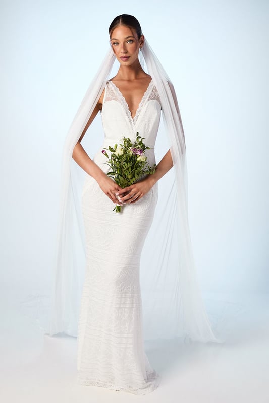 Elegant plunge neckline with delicate lace detailing for a romantic touch
        Figure-flattering fishtail silhouette that gracefully contours the body
        Stunning open back design creating a captivating rear view
        Lightweight chiffon fabric offering comfortable movement throughout your special day
        Floor-length design with subtle train detail for a dramatic entrance
    
    Make a breathtaking statement on your wedding day with this enchanting boho fishtail gown. The sophisticated plunge neckline frames your dcolletage beautifully, whilst the expertly crafted silhouette skims your curves before gently flaring at the knee for that coveted mermaid effect. The delicate lace detailing adds texture and dimension, creating visual interest without overwhelming the clean lines of the dress. Perfect for both traditional ceremonies and outdoor celebrations, this gown can be styled with a cathedral-length veil as shown for classic bridal elegance, or with fresh floral accessories for a more bohemian aesthetic. Complete your bridal ensemble with minimal jewellery to maintain focus on the gown's exquisite design, and pair with strappy heels for added height and poise as you glide down the aisle.