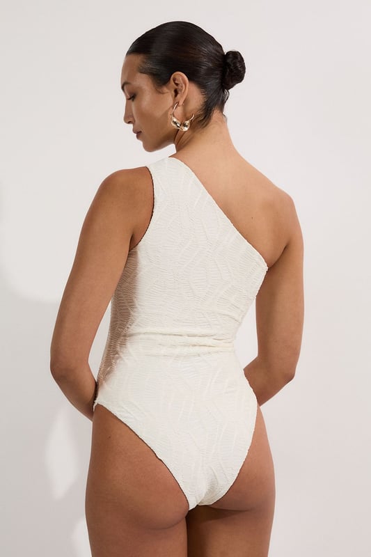 Make a statement poolside in this Karen Millen textured swimsuit that combines sophistication with modern design elements. The asymmetric one-shoulder neckline creates a dramatic focal point, enhanced by the luxe metallic hardware detail. The distinctive cut-out adds intrigue while maintaining a flattering shape. The textured fabric offers visual interest and dimension that catches the light beautifully as you move. Style with oversized sunglasses and gold-toned jewellery for lounging by the pool, or pair with a flowing maxi skirt and sandals for beachside dining. This versatile piece transitions effortlessly from daytime swimming to sunset cocktails when worn as a bodysuit with wide-leg trousers. Perfect for your summer holiday wardrobe, this swimsuit delivers both style and substance for confident beach days.   Striking one-shoulder design with metallic hardware detail Textured fabric creates a sophisticated dimensional effect Strategic cut-out detail adds a contemporary twist Fully lined for comfort and confidence Sculpting fit that flatters the silhouette