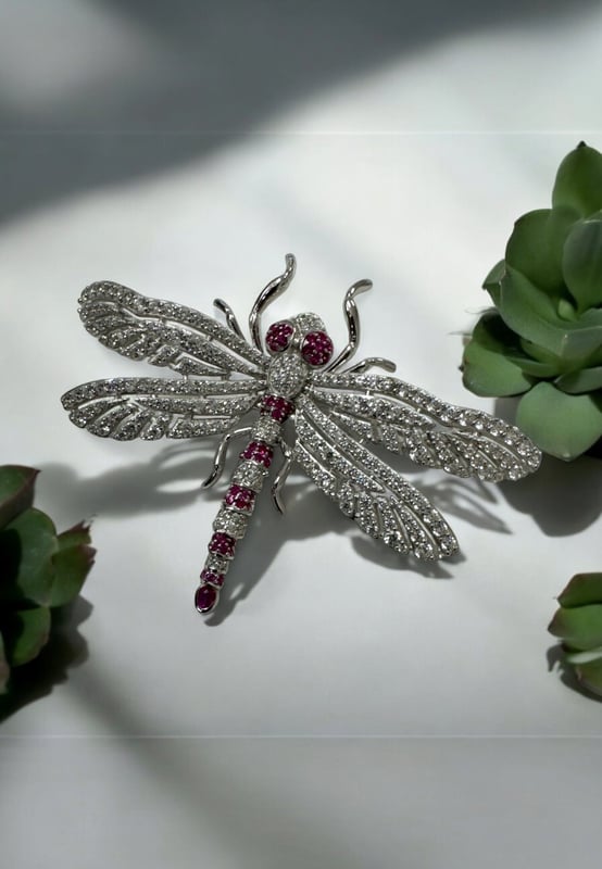 Dragonfly Brooch Silver Ruby Cz The Dragonfly Brooch is a masterfully crafted piece made from high-quality 925 sterling silver. This exquisite brooch captures the delicate beauty of a dragonfly, adorned with a dazzling array of ruby pink and white zirconia. The body of the dragonfly features alternating stripes of pink and white, along with pink eyes, creating a stunning visual contrast that is sure to catch the eye.The wings are intricately designed with a lace-like pattern, encrusted with sparkling white zirconia that shimmer with every movement. The tail of the dragonfly is elegantly finished with a larger ruby-coloured zirconia, making this brooch a true statement piece. Measuring 7.0cm in width, 5.0cm in length, and weighing a comfortable 12.7 grams, this brooch is perfect for adding a touch of glamour to any outfit.Dragonflies hold a special place in jewellery, symbolizing transformation, adaptability, and a deep connection to nature. They remind us to embrace change and live life to the fullest, making this brooch not just a beautiful accessory but a meaningful piece of art.Perfect for nature enthusiasts, lovers of fine craftsmanship, and anyone who appreciates unique and symbolic jewellery. Add the Dragonfly Brooch Silver Ruby Cz to your collection today and let its beauty and symbolism inspire you.