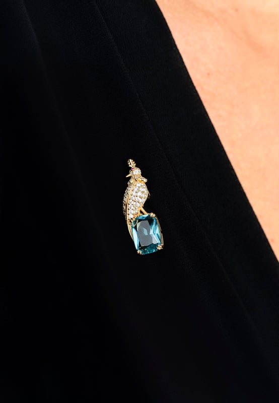 Crested Love Bird Brooch London Blue Topaz Gold Your brooch is a statement of elegance, inspired by the quiet majesty of nature. Designed in the image of a regal crested bird, this handcrafted piece is forged in 925 sterling silver and richly plated with 18ct gold for a luxurious finish that gleams with timeless beauty.Nestled beneath the zirconia-encrusted bird, a lab-created London blue topaz—cut to a refined rectangular silhouette—adds a deep, oceanic sparkle that captivates in every light. At 3.8 cm long, 1.4cm wide and just 7.0 grams in weight, your brooch wears with comfortable grace on any lapel, scarf, or dress.Whether you're accenting a formal look or adding a touch of opulence to your everyday style, your brooch offers a wearable blend of art, craftsmanship, and storytelling. This is more than an accessory—it’s a piece that speaks.