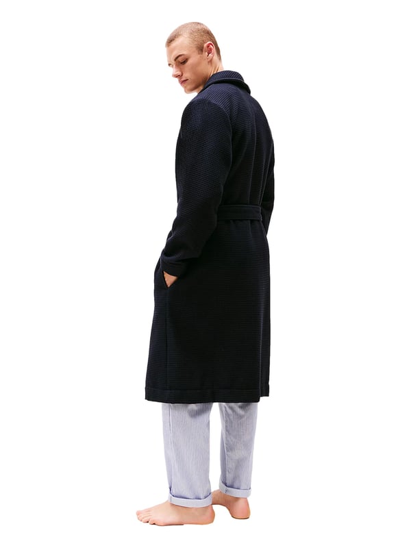 Tommy Hilfiger Bademantel Dieser Herren-Bademantel überzeugt mit stilvoller Waffelstruktur und angenehm zu tragendem Gefühl. Der klassische Schalkragen verleiht ihm eine elegante Note und lädt zum Einkuscheln ein. Mit dem Bindegürtel lässt sich die Passform individuell anpassen – ganz nach deinem Komfort. Zwei aufgesetzte Taschen vorne bieten Platz für kleine Essentials oder einfach warme Hände. Leicht überschnittene Schultern sorgen für eine entspannte Silhouette und zusätzliche Bewegungsfreiheit. Das Label-Necktape im Nackenbereich unterstreicht die hochwertige Verarbeitung. Eine dezente Logo-Stickerei auf der Brust setzt ein geschmackvolles Markenstatement. Der Stoff ist leicht, aber dennoch wärmend – ideal für gemütliche Morgen oder entspannte Abende. Ob nach der Dusche oder beim Frühstück auf dem Balkon, dieser Bademantel macht jede Auszeit noch angenehmer. Für Männer, die sich stilvoll entspannen wollen.