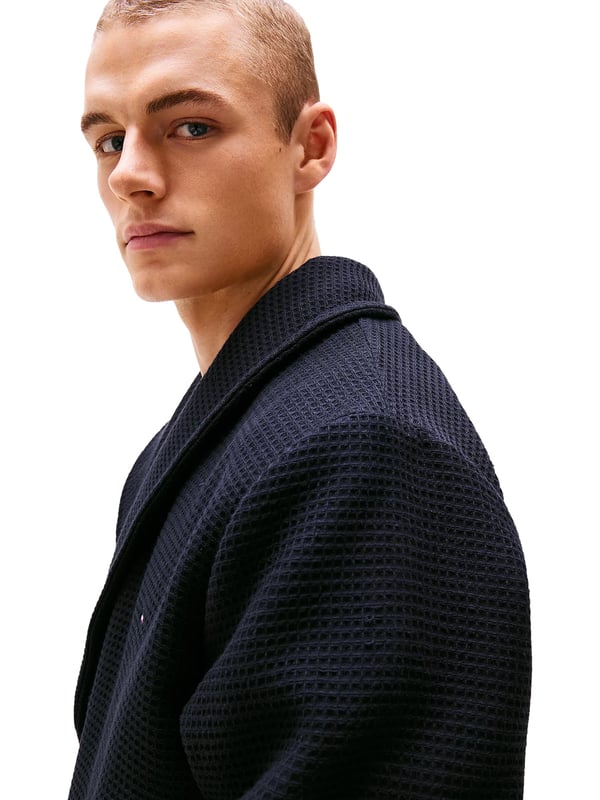 Tommy Hilfiger Bademantel Dieser Herren-Bademantel überzeugt mit stilvoller Waffelstruktur und angenehm zu tragendem Gefühl. Der klassische Schalkragen verleiht ihm eine elegante Note und lädt zum Einkuscheln ein. Mit dem Bindegürtel lässt sich die Passform individuell anpassen – ganz nach deinem Komfort. Zwei aufgesetzte Taschen vorne bieten Platz für kleine Essentials oder einfach warme Hände. Leicht überschnittene Schultern sorgen für eine entspannte Silhouette und zusätzliche Bewegungsfreiheit. Das Label-Necktape im Nackenbereich unterstreicht die hochwertige Verarbeitung. Eine dezente Logo-Stickerei auf der Brust setzt ein geschmackvolles Markenstatement. Der Stoff ist leicht, aber dennoch wärmend – ideal für gemütliche Morgen oder entspannte Abende. Ob nach der Dusche oder beim Frühstück auf dem Balkon, dieser Bademantel macht jede Auszeit noch angenehmer. Für Männer, die sich stilvoll entspannen wollen.