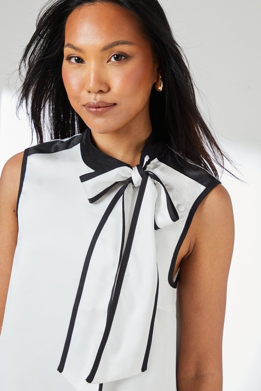 Sleeveless satin top with striking contrast trim along the neckline and armholes
        Elegant tie-neck detail creates a sophisticated bow at the collar
        High neckline offers a refined silhouette perfect for formal settings
        Clean, straight-cut hemline for a polished finish
        Smooth woven fabric with a subtle lustre for an elegant appearance
    
    This Principles sleeveless top combines timeless elegance with contemporary design through its distinctive contrast detailing. The sophisticated tie-neck feature allows for versatile styling-wear it neatly tied for formal occasions or slightly loosened for a more relaxed interpretation. Pair with wide-leg trousers as shown for a chic monochromatic ensemble that transitions seamlessly from office to evening events. For formal occasions, style with a tailored blazer, statement earrings and court shoes. Alternatively, create a smart-casual look by teaming with slim-fit trousers and pointed flats. The clean lines and classic contrast colouring make this top an excellent foundation piece for multiple outfits, whether attending a wedding reception, business meeting, or sophisticated dinner engagement.