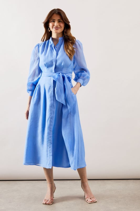 Elegant textured organza midi shirt dress with collarless neckline for a sophisticated silhouette
        Voluminous balloon sleeves with gathered cuffs create a dramatic yet refined look
        Flattering waist tie belt accentuates the figure while allowing for customised fit
        Practical side pockets discreetly integrated into the design
        Midi length offers versatility for both daytime and evening occasions
    
    This striking textured organza shirt dress combines contemporary elegance with timeless sophistication. The collarless design and button-through front create a clean, refined aesthetic, while the sumptuous balloon sleeves add a touch of drama to your ensemble. Cinch the included waist tie to create a beautifully defined silhouette that flatters all figures. Perfect for weddings and formal gatherings, style with delicate strappy heels as shown and statement earrings for a polished look. For race day events, complement with a fascinator and clutch bag. The versatile midi length works beautifully for graduation ceremonies or elegant garden parties. This Wallis piece transitions effortlessly from day to evening, making it a valuable addition to your occasion wardrobe that will serve you through multiple seasons with different accessories.
