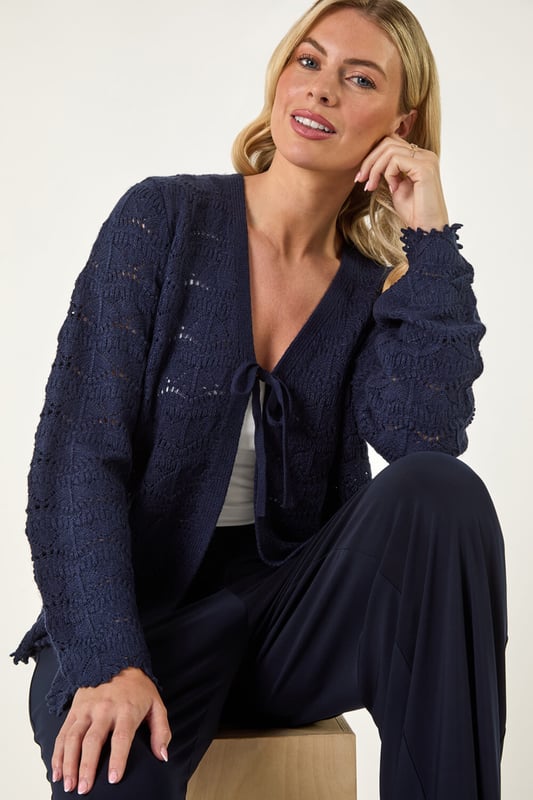 Petite Knitted Tie Cardigan from Roman. Elevate your knitwear collection with this charming v-neck cardigan. Crafted from a knitted fabric, this edge to edge cardigan features a unique tie fastening at the bust for added detail. The scallop style hem and pointelle detail along with the long sleeves make this cardigan a standout piece. Pair with jeans and sandals for a laid-back and stylish look this Spring/Summer season!